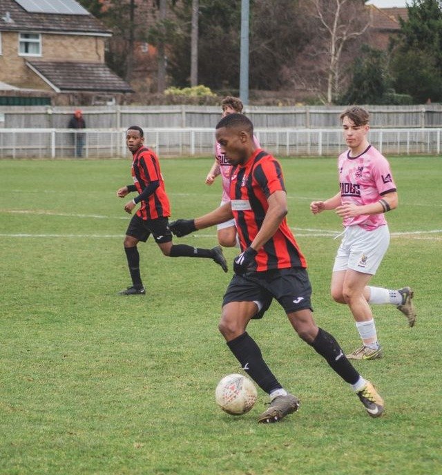 A soccer player in a black and red striped jersey dribbles the ball downfield while two other players follow.