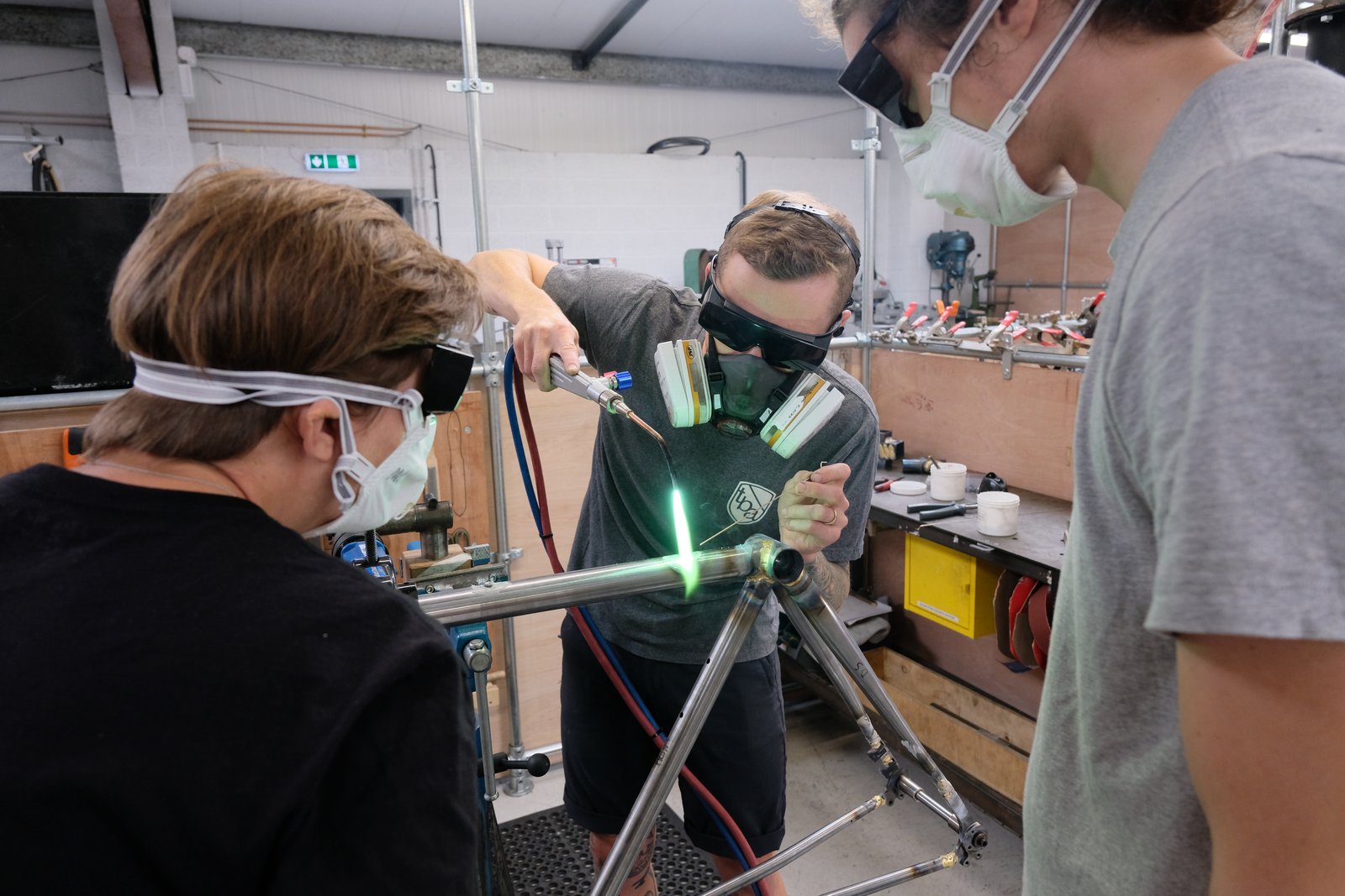 Three people watch a demo of someone building a bike frame