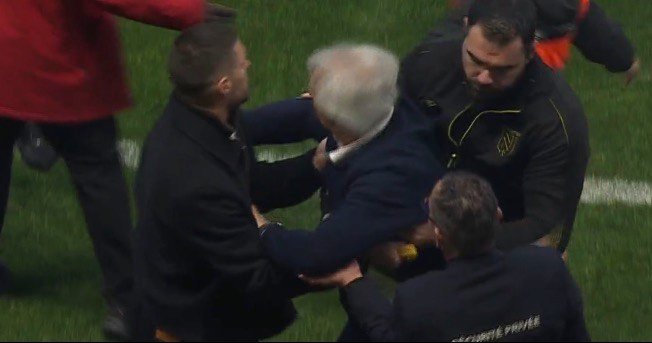 Several men in jackets physically restraining a white-haired man on a grassy field.