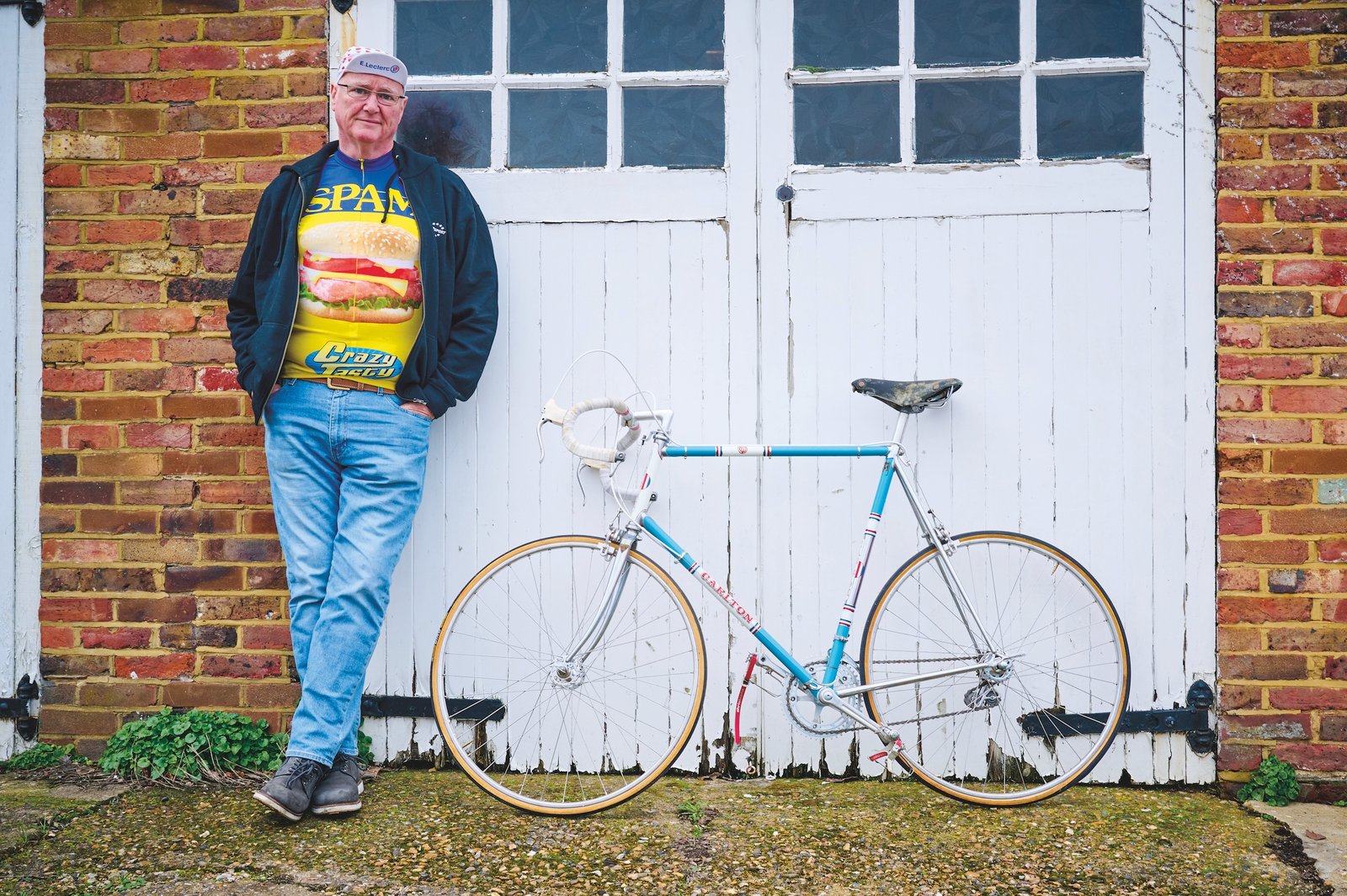 Carlton Kirby standing leaning on his white garage doors alongside his vintage racing bike